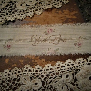 May include: A beige fabric ribbon with a frayed edge and a floral pattern. The ribbon has the words "With Love" embroidered in brown thread.