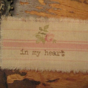 Puede incluir: Una tela beige y rosa con un borde deshilachado, que presenta un pequeño diseño floral bordado y el texto "in my heart".