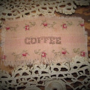 May include: A pink fabric with a frayed edge, embroidered with a floral pattern and the word "COFFEE" in brown thread.