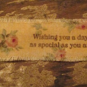 May include: A rectangular burlap ribbon with frayed edges. The ribbon is a light yellow color and features a floral pattern with pink roses and green leaves. Text on the ribbon reads, "Wishing you a day as special as you are."