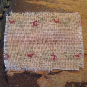 May include: A small, beige fabric square with a frayed edge. The fabric has a delicate floral pattern with small red flowers. The word "believe" is embroidered in brown thread in the center of the square.