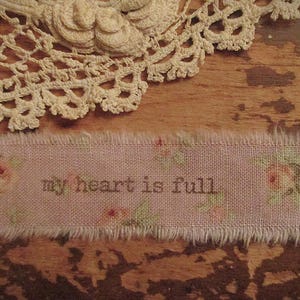 Op de afbeelding: Een close-up van een roze stoffen strook met de bruine tekst "my heart is full". De strook heeft een bloemenpatroon en gerafelde randen. Op de achtergrond een crèmekleurig kanten kleedje.
