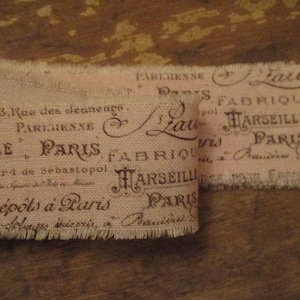 Puede incluir: Dos tiras de cinta de tela de estilo vintage con bordes deshilachados. La cinta es de color beige claro con texto marrón oscuro con las palabras "París" y "Marsella". La cinta probablemente se utiliza para manualidades o fines decorativos.