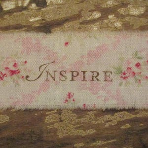 May include: A rectangular fabric tag with the word "INSPIRE" in brown script. The tag has a frayed edge and a floral pattern in pink and green on a cream background. The tag is on a textured brown surface.