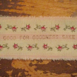 May include: A beige fabric ribbon with pink flowers and the text "Be good for goodness sake" embroidered in brown thread.