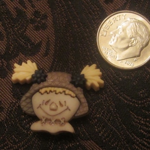 Può includere: Una statuetta di spaventapasseri marrone e beige con un cappello di paglia e occhi neri. Lo spaventapasseri sorride e ha un corpo di stoffa marrone e beige.