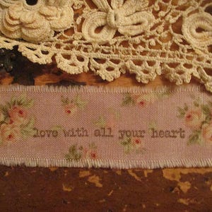 Peut inclure: Une bande de tissu rose avec un motif floral et le texte "love with all your heart". La bande a un bord effiloché et est partiellement recouverte d'une garniture en dentelle crème. Le fond est une surface en bois foncé.