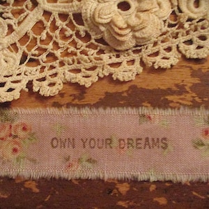 Peut inclure: Ruban rose avec l'inscription « OWN YOUR DREAMS » et des motifs floraux. Le ruban a un bord effiloché et est posé sur une surface en bois. Une nappe en dentelle crème et un petit groupe de perles sont également visibles.