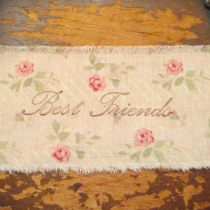 May include: A white fabric with a floral pattern and the words "Best Friends" written in cursive. The fabric has a frayed edge.