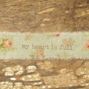 May include: A rectangular fabric strip with frayed edges, featuring the text "my heart is full" in a light brown colour. The strip is decorated with pink roses and green leaves on a light green background.