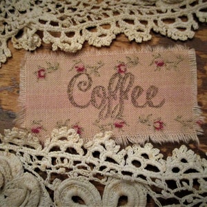 May include: A rectangular piece of fabric with the word "Coffee" in cursive script, surrounded by small floral accents. The fabric is framed by delicate, cream-colored lace, creating a vintage aesthetic.