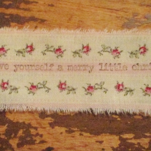 May include: A rectangular fabric banner with frayed edges. The banner is cream-colored with a pink stripe in the center. It is decorated with small red roses and green leaves. The text "have yourself a merry little christmas" is embroidered in the center.