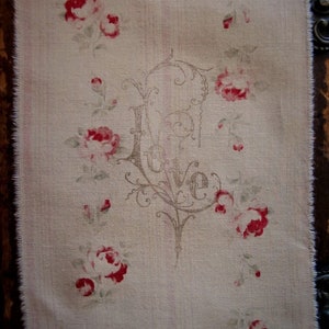 以下が含まれることがあります： フリンジの付いたベージュの生地に、茶色の装飾的な文字で「Love」という言葉が書かれています。生地には、小さな赤いバラの模様が施されています。