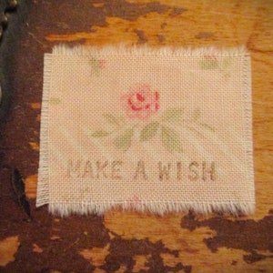Puede incluir: Un parche de tela rectangular con bordes deshilachados, con un estampado floral descolorido y el texto "MAKE A WISH" en fuente marrón. El diseño incluye una rosa rosa y hojas verdes.