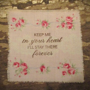 May include: A square, pale pink fabric with a frayed edge, adorned with floral patterns and the text "KEEP ME in your heart I'LL STAY THERE forever" in brown. The design has a vintage, romantic feel.