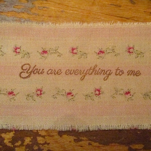 May include: A rectangular piece of fabric with the phrase "You are everything to me" embroidered in brown. The fabric is light pink with a floral border of red roses and green leaves. The edges are frayed.