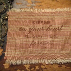 May include: A peach-coloured fabric rectangle with a frayed edge, inscribed with the words "KEEP ME in your heart I'LL STAY THERE forever" in brown script. The fabric has a cream striped pattern.