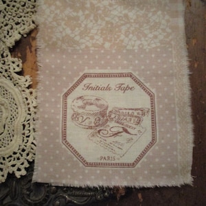 Pode incluir: Um tecido rosa com bolinhas brancas e um rótulo octogonal marrom que diz "Initials Tape - PARIS" com uma ilustração vintage de carretéis de linha e uma agulha.