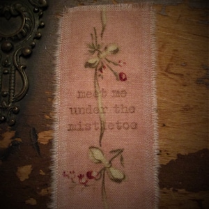 May include: A rectangular piece of pink fabric with frayed edges, featuring embroidered text that reads "meet me under the mistletoe." A delicate floral design with red berries and green leaves is embroidered above and below the text.