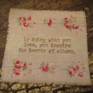 Puede incluir: Una tela blanca con un borde deshilachado presenta una cita bordada en negro: "Haciendo lo que amas, inspiras los corazones de los demás." La tela está decorada con un patrón floral de rosas rosas.