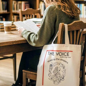 May include: A white tote bag with the text "THE VOICE" and "Wake up the sleepers" in red and black. The bag features a line drawing of a woman's face and floral elements. The bag is hanging on a wooden chair in a library.