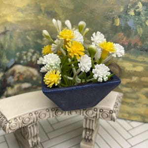 May include: A miniature arrangement of yellow and white flowers in a square, navy blue ceramic pot. The pot sits on a small, decorative, off-white bench with ornate detailing. The background features a blurred painting with green and yellow hues.
