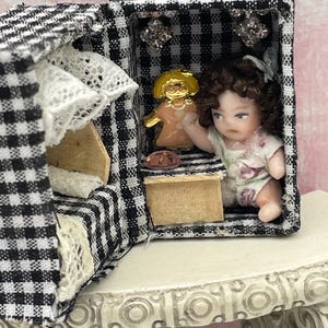 May include: A miniature room box lined with black and white gingham fabric containing a small doll with brown curly hair wearing a floral outfit. Inside, a gold-toned figurine, a pink circular accent, and white lace trim are visible.