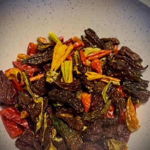 May include: A white bowl filled with a variety of dried peppers. The peppers are red, orange, yellow, and green. Some of the peppers are whole, while others are broken into pieces.