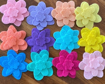 Könnte beinhalten: Eine Sammlung bunter Filzblumen-Verzierungen in verschiedenen Farbtönen, darunter Rosa, Lila, Orange, Blau und Gelb. Jede Blume hat mehrere Schichten, wodurch ein dreidimensionaler Effekt entsteht. Ideal für DIY-Projekte.