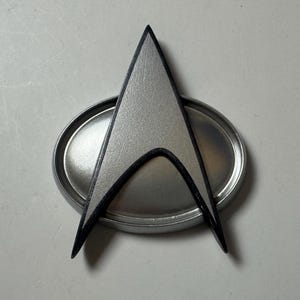 Puede incluir: Una insignia de mando de la Flota Estelar plateada con un contorno negro, sobre una base ovalada. La insignia presenta un diseño de punta de flecha estilizada, un símbolo de la franquicia de ciencia ficción Star Trek.