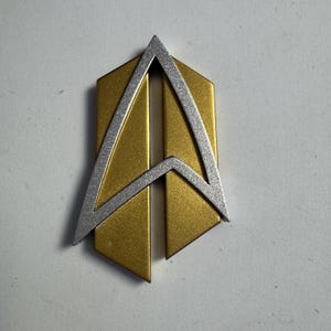 May include: A gold and silver Starfleet insignia pin. The pin has a gold, shield-shaped base with a silver delta-shaped emblem across the centre. The design is a stylized representation of a star and arrow.