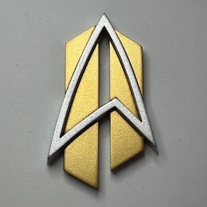 May include: A metallic emblem featuring a textured gold-coloured vertical rectangular base with a pointed bottom edge. Overlaid on the gold base is a silver-coloured, angular, arrow-like frame with a sharp top point and two downward-extending legs.