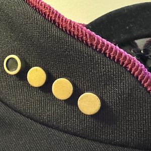 May include: Close-up of a black garment with gold-colored buttons. The edge is trimmed with a magenta-colored decorative trim. The garment appears to be made of a textured fabric.