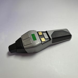 May include: A silver and black prop device with a rectangular shape. It features a green display, gold-colored buttons, and a textured grip. The device is likely a replica or a prop from a science fiction series.