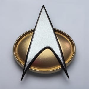 Puede incluir: Una insignia de comunicador de Star Trek que presenta un emblema plateado en forma de delta con un borde negro, montado sobre una base ovalada lisa de color dorado. El diseño consta de una parte superior puntiaguda y dos lados curvos.