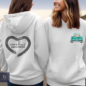 Puede incluir: Dos sudaderas con capucha blancas. Una sudadera con capucha tiene un corazón negro con un diseño de banda de rodadura de neumáticos y el texto "Tengo un corazón como un camión". La otra sudadera con capucha tiene una camioneta azul turquesa con flores en la parte trasera y el texto "Jenny Wilson".