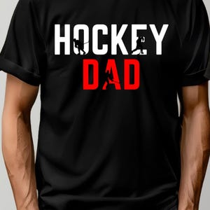 Puede incluir: Camiseta negra con texto blanco en mayúsculas que dice "HOCKEY DAD". El texto está superpuesto a una silueta de jugadores de hockey.