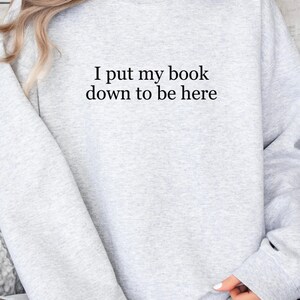 Op de afbeelding: Grijs sweatshirt met de tekst "I put my book down to be here" gedrukt op de voorkant.