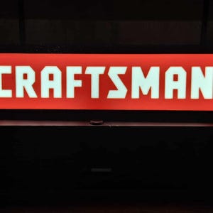 May include: A red rectangular sign with the word "CRAFTSMAN" in white block letters. The sign is illuminated and set against a dark background. The image suggests a focus on tools or hardware.