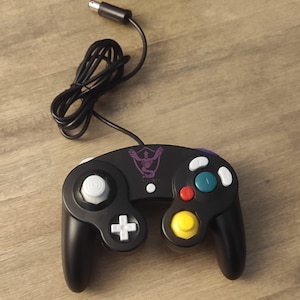 Puede incluir: Un mando de GameCube negro con un diseño morado y blanco. El mando tiene un stick C blanco, botones amarillos y rojos, y un logotipo morado y blanco en la parte delantera.