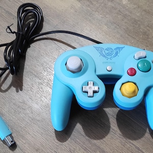 Peut inclure: Une manette GameCube bleue avec un logo de crâne et d'ailes. La manette a un bouton jaune, rouge et vert. La manette est connectée à un cordon noir.