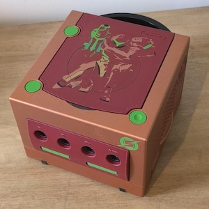 Puede incluir: Una consola Nintendo GameCube pintada a medida en color cobre con una parte superior roja. La parte superior presenta una silueta de Samus Aran de la serie de videojuegos Metroid. La consola tiene botones verdes y una unidad de disco negra.