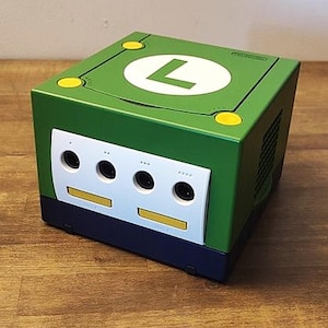Può includere: Una console Nintendo GameCube verde personalizzata con un grande cerchio bianco e una "L" verde sulla parte superiore. La console ha un pannello frontale bianco con porte per controller nere e dettagli gialli. La base è blu scuro.