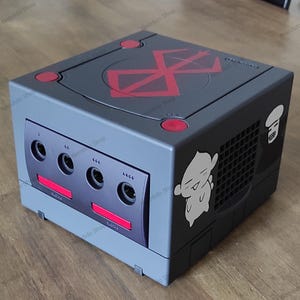 Può includere: Una console Nintendo GameCube grigia personalizzata con dettagli rossi e adesivi. La parte superiore presenta un simbolo rosso, e il lato ha un adesivo di un personaggio dei cartoni animati bianco. La parte anteriore ha quattro porte controller e un pulsante di accensione rosso.