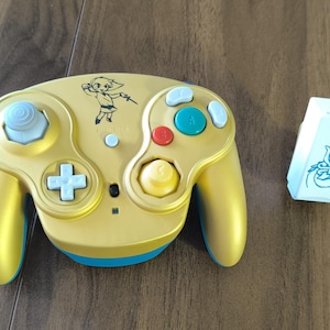 May include: A gold-colored video game controller with blue and white buttons and a blue base. The controller has a cartoon graphic of a character holding a sword. A white rectangular device with a blue graphic is next to the controller.