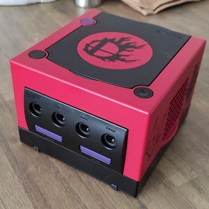 Puede incluir: Una consola Nintendo GameCube roja con una parte superior negra y un logotipo de Super Smash Bros. La consola tiene cuatro ranuras para juegos y un botón morado en la parte superior.