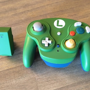 Peut inclure: Manette de jeu Nintendo GameCube verte et bleue avec un 'L' blanc sur le devant. La manette a un stick C jaune, un bouton plus blanc et un stick analogique blanc. La manette est connectée à un récepteur vert.
