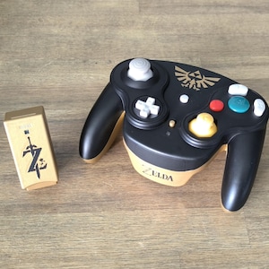 Może przedstawiać: Czarno-złoty kontroler Nintendo GameCube z logo Zeldy i dopasowanym złotym odbiornikiem bezprzewodowym. Kontroler ma biały analogowy drążek, czarny krzyż kierunkowy i żółty drążek C.