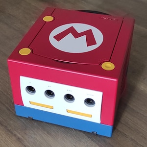 Op de afbeelding: Een rode Nintendo GameCube console met een wit Mario-logo op de bovenkant. De console heeft vier controllerpoorten en een blauwe basis.