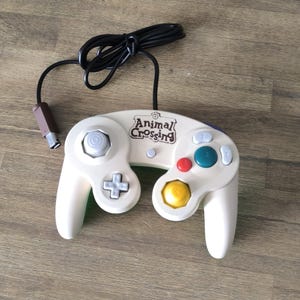 Puede incluir: Mando de GameCube de Animal Crossing de color crema con cable negro. El mando tiene una cruceta, una palanca analógica y varios botones de colores, incluyendo rojo, azul y amarillo. El logotipo de Animal Crossing es visible.